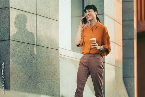 Young professional woman talking on phone and holding coffee outside modern office building