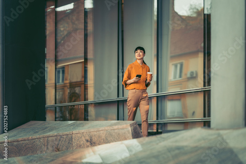 Young professional woman walking outside modern office building with smartphone and coffee cup