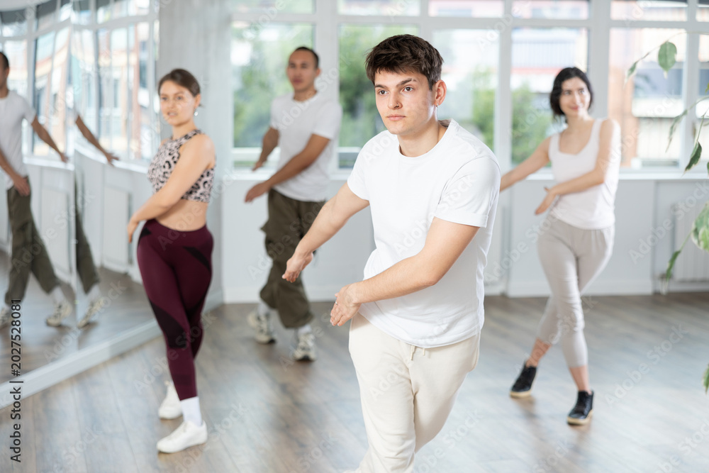 Fototapeta premium Enthusiastic sporty smiling Hispanic guy energizing group of young people during dance fitness class in well-lit mirrored studio
