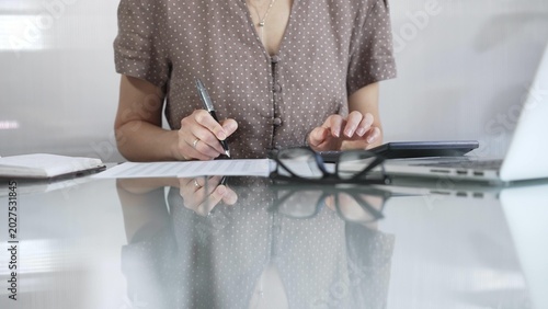Quadro su tela Businesswoman accountant, wearing brawn dotted blouse, is using a calculator to crunch numbers and jotting down meticulous notes, at sleek glass desk in a contemporary office