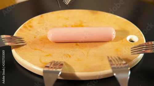 Close-up view of a person slicing a single sausage into several small pieces with a knife and fork on a wooden cutting board, with many forks ready to grab a portion of the food