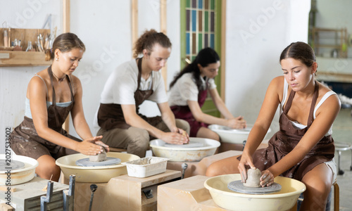 Photos Interested young woman in pinafore making clay pot by potter's wheel in art stud