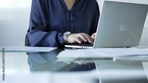Bild auf Leinwand Businesswoman in blue blouse typing on laptop at glass desk while sitting in bright office setting, close-up vie