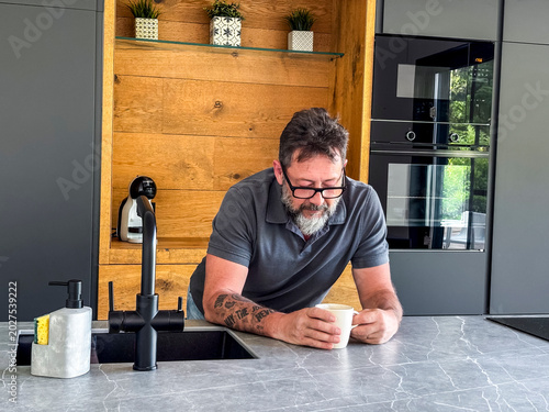 Foto Bearded man with tattoo enjoying coffee in modern kitchen leaning on marble isla