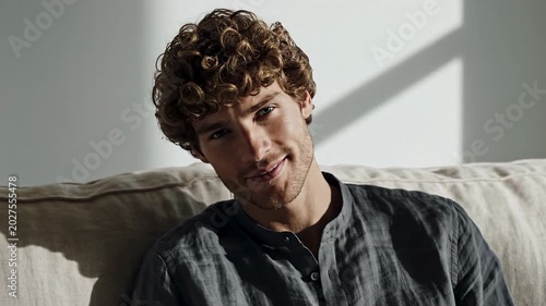 Young man with curly hair relaxing on a sofa in a sunlit room, calm friendly expression and casual clothing, lifestyle portrait with natural window light