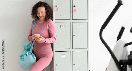 Mature woman with sports bag using mobile phone near locker at gym