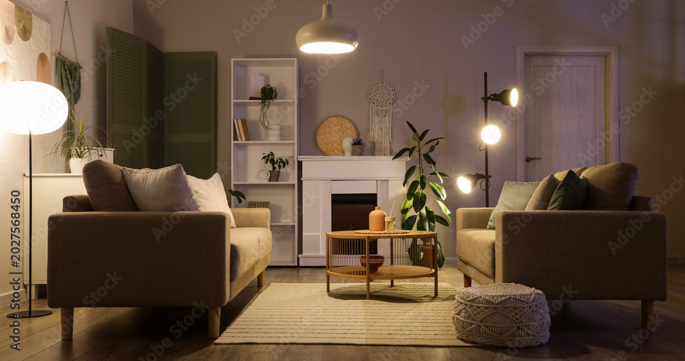Fototapeta premium Interior of stylish living room with grey sofas, coffee table, fireplace and glowing lamps at evening