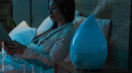 Obraz na płótnie Air humidifier on table against woman reading book in bedroom at night, closeup