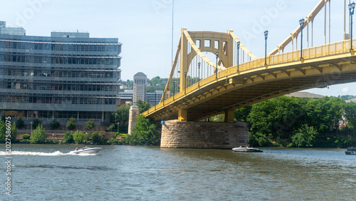 A scenic, low-angle shot of the iconic yellow Roberto Clemente Bridge (Sixth Street Bridge) stretching across the Allegheny River in downtown Pittsburgh, Pennsylvania. On the left, a modern glass-faca
