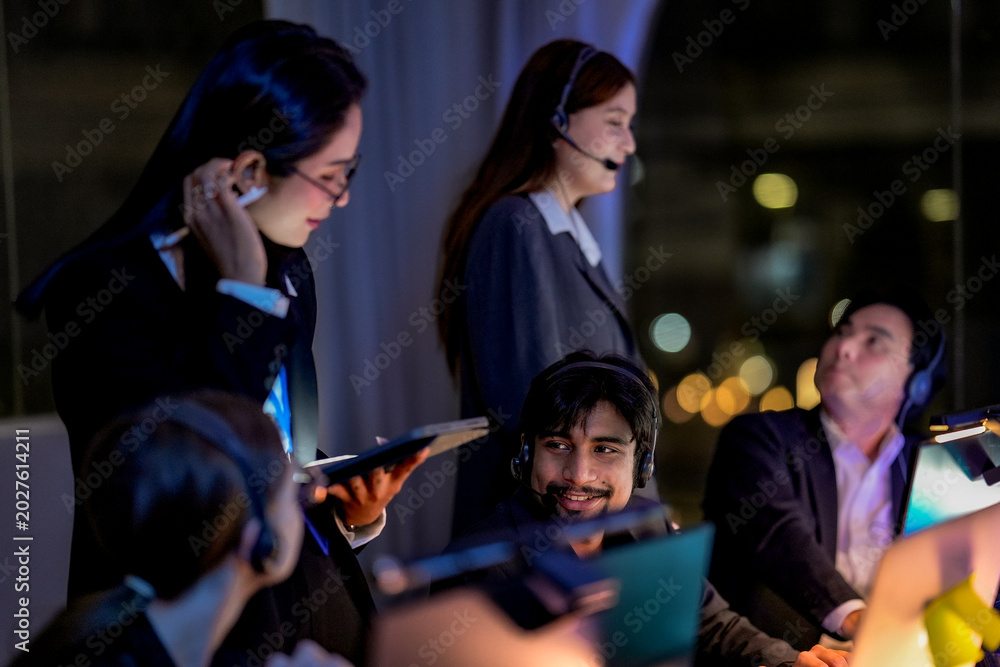 Fototapeta premium Female supervisor helping a male customer service agent during night shift. Diverse call center team working overtime in a modern dark office with beautiful city lights.