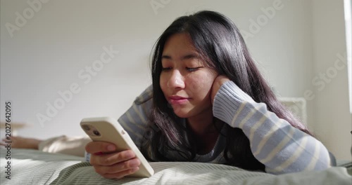 Asian Teenage girl lies on neatly made bed,  Soft morning light from , propped on elbows face, using phone