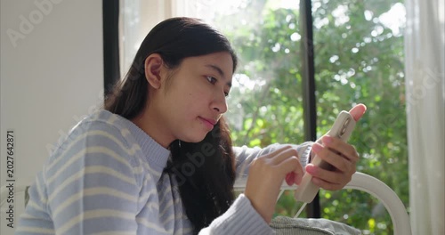 Asian teenage girl sits near a large window, using smartphone, Sunlight filters through sheer curtains in bedroom, using phone, Side view
