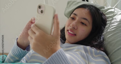 Asian teenage girl lying near on bed, using smartphone, Sunlight filters through sheer curtains in bedroom