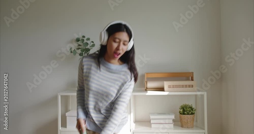 Asian woman girl in a striped sweater dances with a mop as a microphone in a bright minimalist home, wearing white headphones