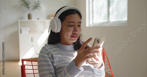 Asian teenage girl sits indoor listen to music, wearing white headphones and a light blue striped sweater, using smartphone