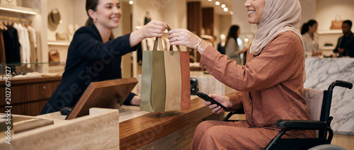 Close-up of Muslim woman in wheelchair receiving paper shopping bag from seller in a luxury boutique, holding smartphone after paying for her purchase - Wide panoramic banner or website header