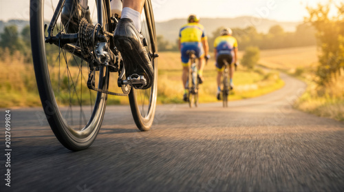 Closeup of a cyclists feet pedaling on a road bike with two other cyclists riding ahead on a scenic country road during sunset