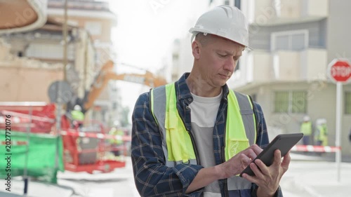 Man builder in hardhat and hi vis vest taps tablet while checking plans on a street construction site; focused inspection.