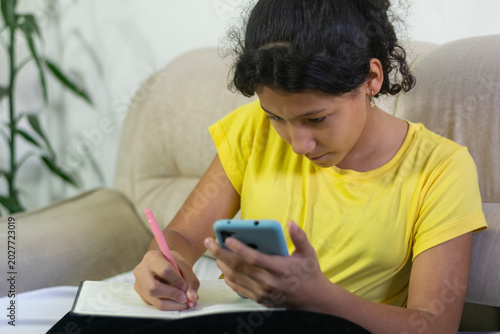 Latina preteen girl copying notes from smartphone into notebook while sitting on sofa at home