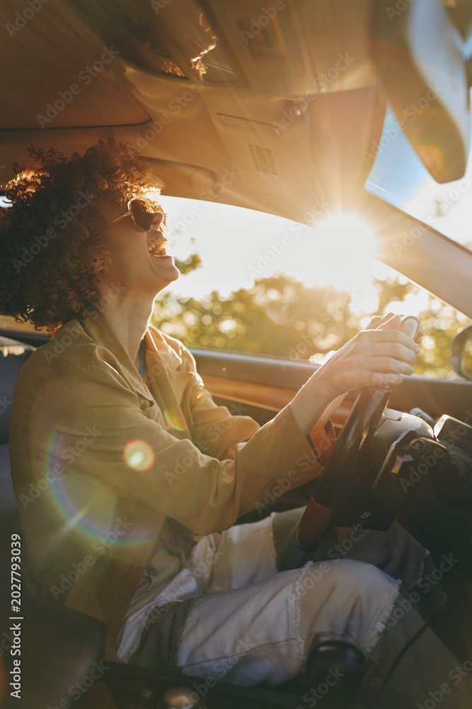 Fototapeta premium Smiling woman with curly hair driving a car during golden hour. Sunlight fills the cabin with a warm glow, creating a relaxed lifestyle moment focused on travel and self care.