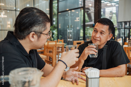 Картината върху платно Two Filipino men sit across from each other in a Southeast Asian bar or restaurant, holding glasses of water during conversation