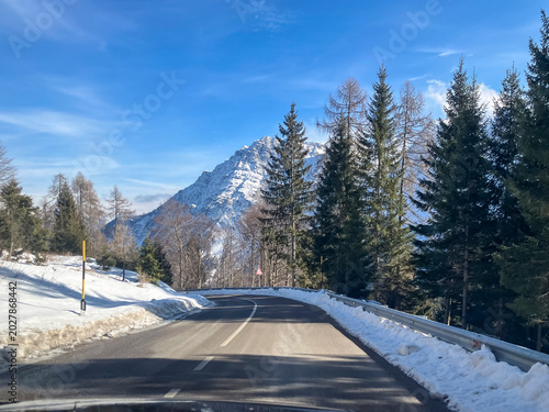 Snowy mountain road winding through a winter forest toward the alpine mountains under a cold blue sky,Italy
