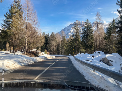 Snowy mountain road leading through a forest toward an alpine ski resort under a cold winter sky,Italy