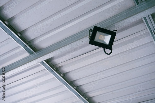 Industrial spotlight hangs from metal beams. White corrugated ceiling reflects bright, cool light. Metal framework supports modern lighting. Ideal for warehouses, factories, or industrial spaces