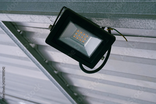 Industrial spotlight hangs from metal beams. White corrugated ceiling reflects bright, cool light. Metal framework supports modern lighting. Ideal for warehouses, factories, or industrial spaces