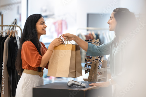 Photography Smiling Brazilian female customer receives shopping bags from boutique employee at checkout counter