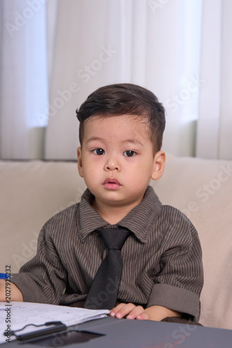 A 2-year-old little boy sitting on the sofa looking directly at the camera while holding a pen