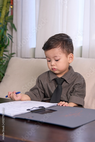 A 2-year-old toddler boy sitting at the table holding a pen and looking down at the paper