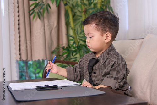 A 2-year-old baby boy sitting at the table holding a blue pen over the clipboard