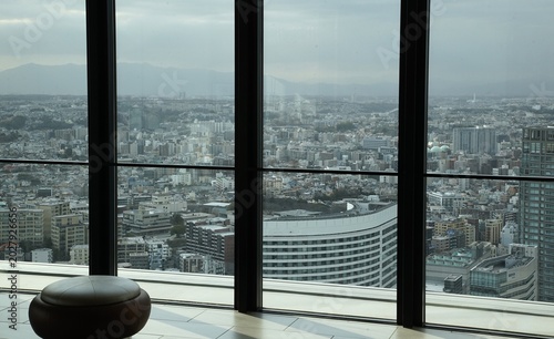 Panoramic urban cityscape viewed through tall floor? to? ceiling windows from an indoor observation area with a round stool
