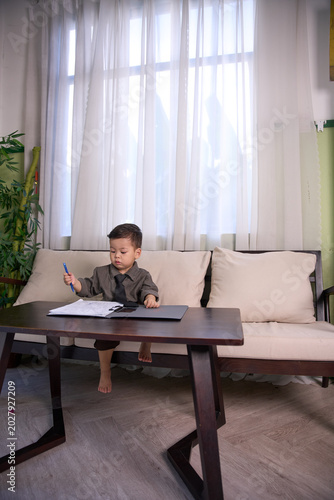 A 2-year-old little boy sitting barefoot at a wooden table with clipboard in front of him