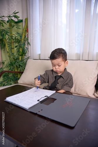 A 2-year-old baby boy sitting at the table writing with a blue pen on a clipboard
