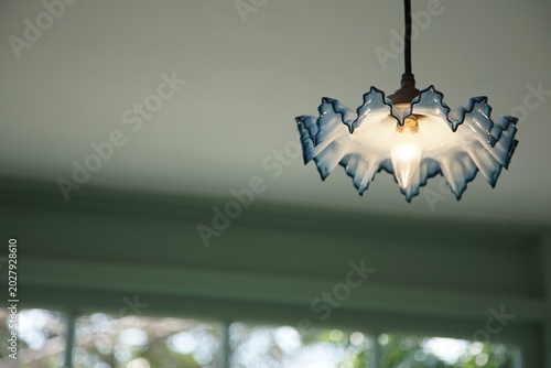 Ceiling view of a single pendant light with a blue edged decorative glass shade and exposed bulb hanging above blurred window frames
