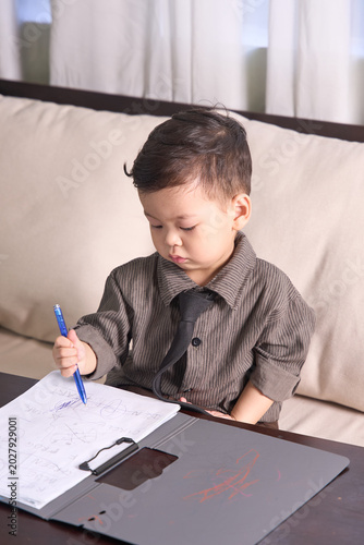 A 2-year-old toddler boy sitting on the sofa writing carefully with a blue pen