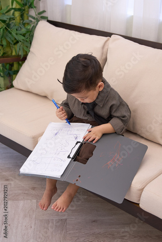 A 2-year-old toddler boy sitting on the sofa looking down and writing on the clipboard