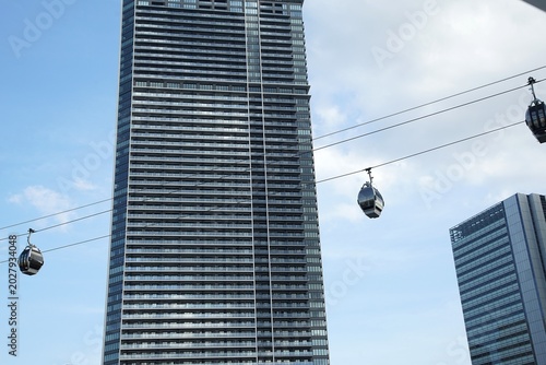 Modern cable cars glide across the sky in front of a tall glass skyscraper, linking parts of a busy waterfront city under soft afternoon clouds