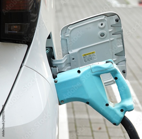 Close-up of a light blue charging plug connected to a white electric car, its open charge port exposed above a paved parking area
