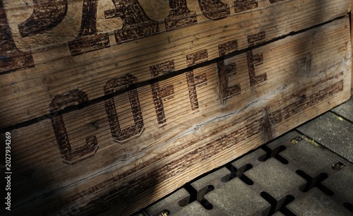 Close-up of a rustic wooden board with the word 'COFFEE' carved and darkened, catching warm sunlight and deep shadows on its textured grain
