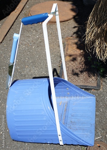 Bright blue outdoor dustpan with metal handle lying on sunlit asphalt beside manhole covers and dry straw shadows