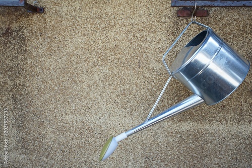 Shiny metal watering can hanging sideways from a rusty hook against a rough beige wall in soft outdoor light