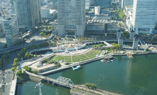 An aerial view of the Minato Mirai district in Yokohama, featuring a docked sailing ship, a grassy park, modern buildings, and a bridge over water
