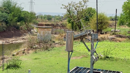 Closeup of solar powered monitoring equipment and antenna installation near a rural riverbank surrounded by green vegetation and dry landscape!