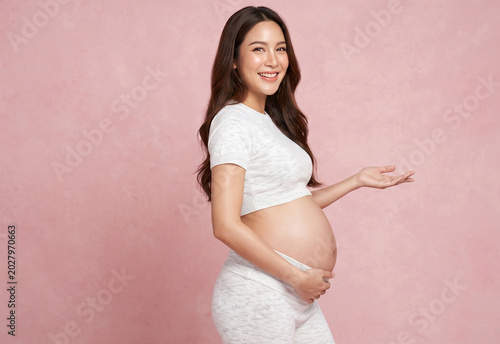 A happy pregnant Asian woman in white activewear smiles warmly while cradling her baby bump and gesturing with an open palm against a textured pink background