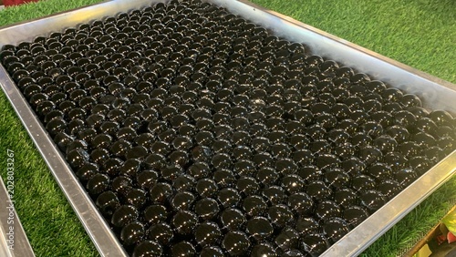 Glossy black tapioca pearls, often referred as giant boba pearls on aluminum tray, infused with flavors like brown sugar or caramel to enhance their taste. commonly used in specialty bubble tea drinks
