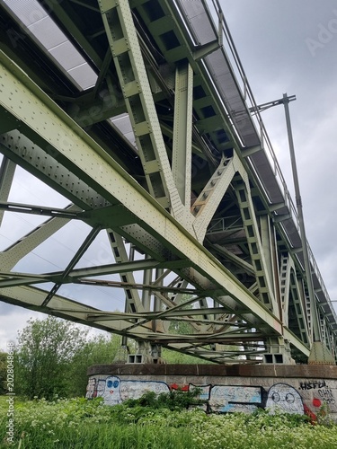 Photography Low angle view of a massive green steel girder railway bridge structure near Arnhem and Oosterbeek, representing Dutch transport infrastructure under an overcast sky