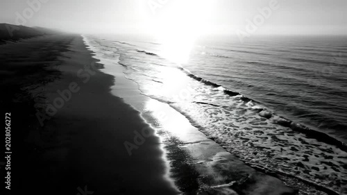 Black and white aerial view of a misty beach with glistening sunlight reflecting on the water at dawn